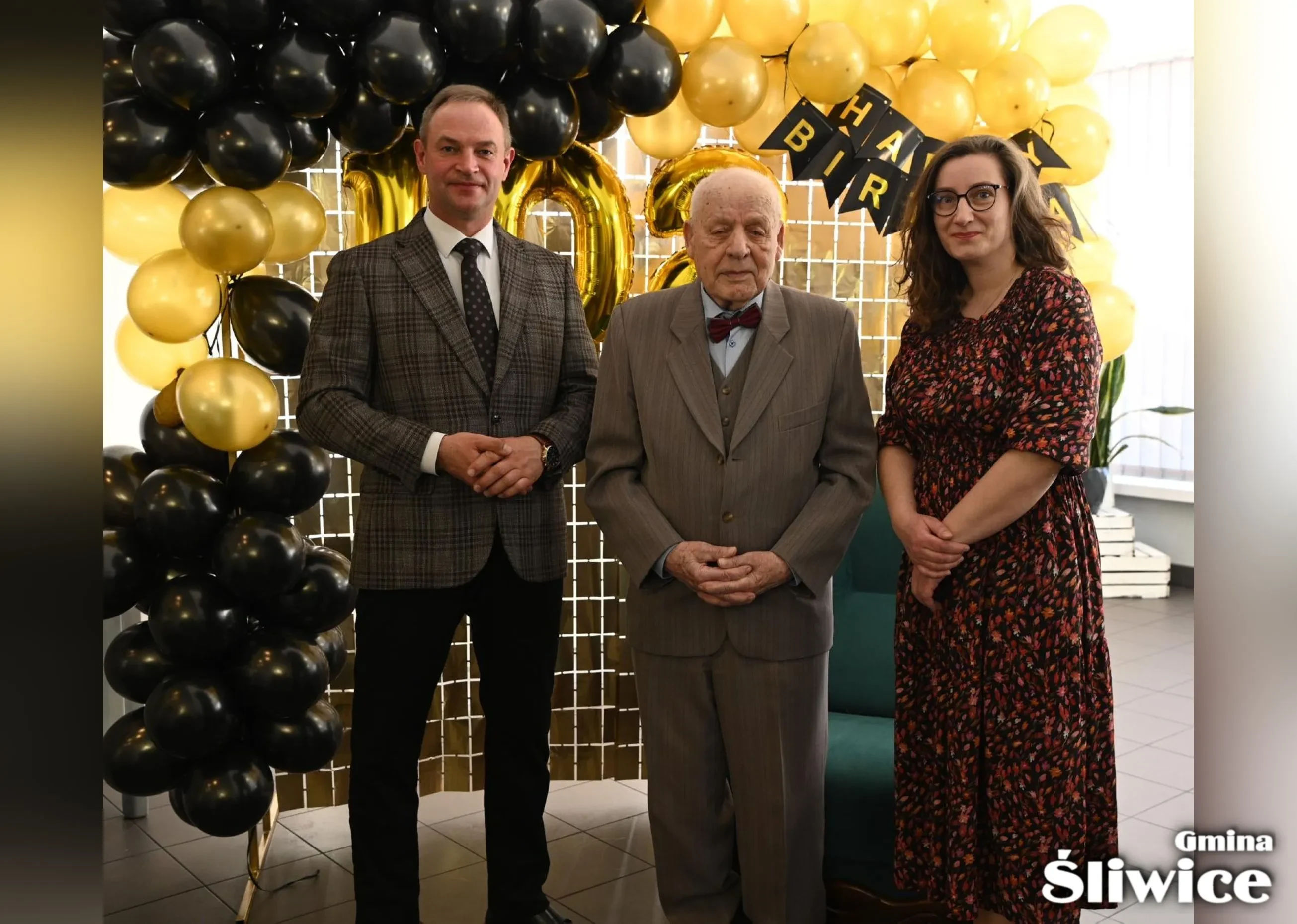 wójt, solenizant i zastępca kierownika usc pozują do zdjęcia, w tle ścianka z balonami